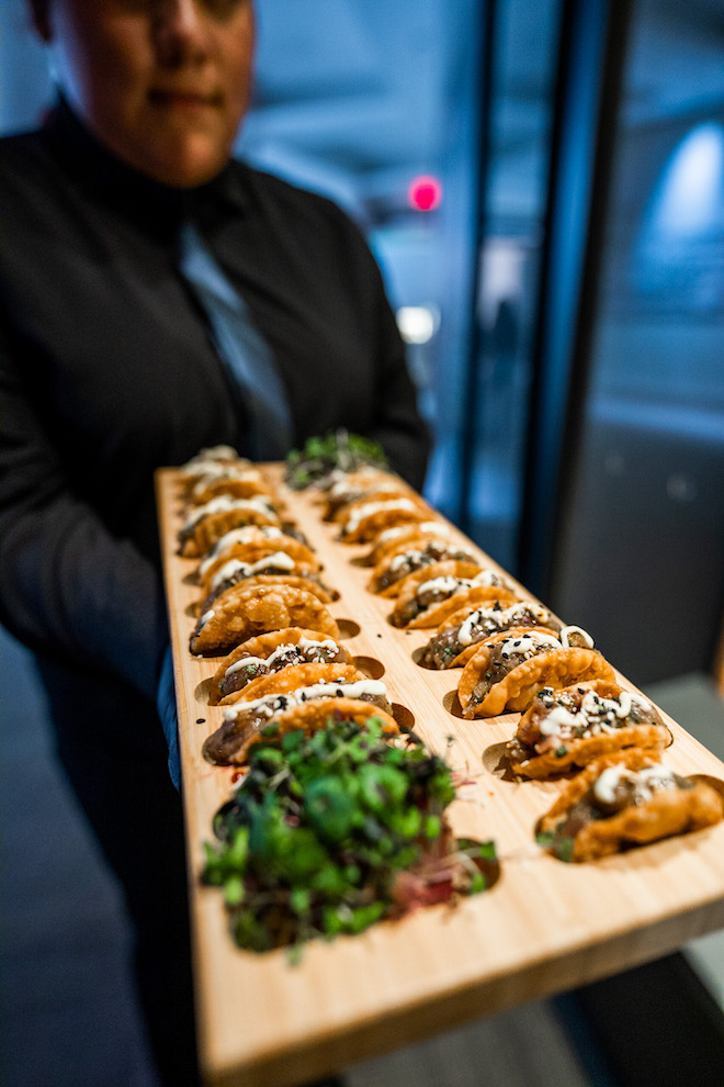 Mini tacos being served by man in suit on a wooden plate.