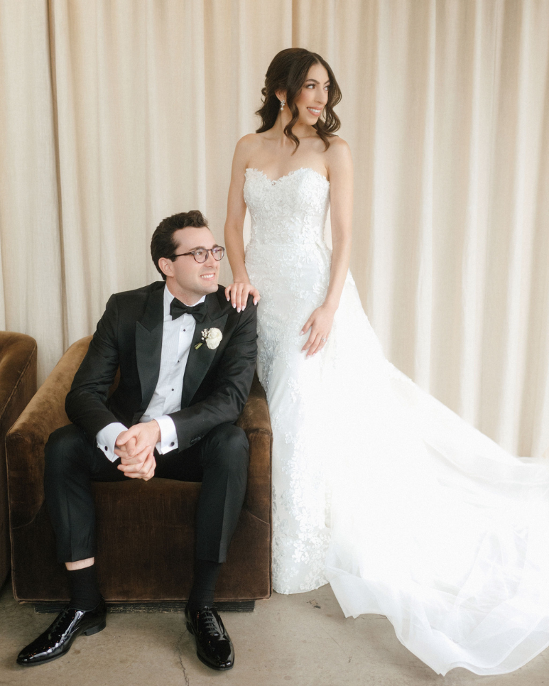Bride and groom smiling and staring to the side while the groom is sat in a chair and the bride has her hand on him standing up. 