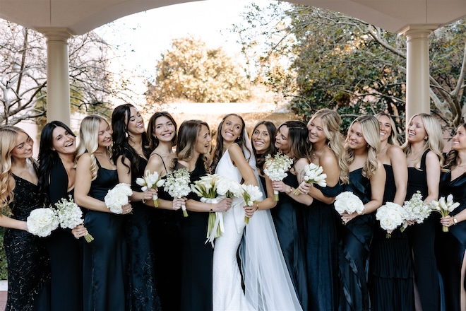 The bride smiling with her bridesmaids wearing black gowns and white bouquets. 
