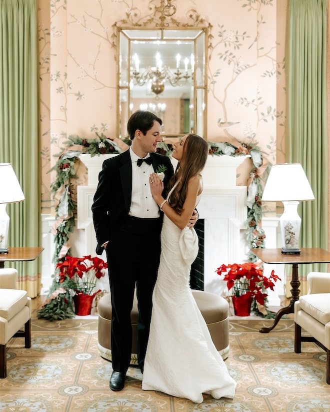 A bride and groom smiling at each other in front of a fireplace decorated with garland and poinsettia. 