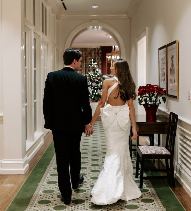 The bride and groom holding hands walking down a hallway with a Christmas tree in front of them. 