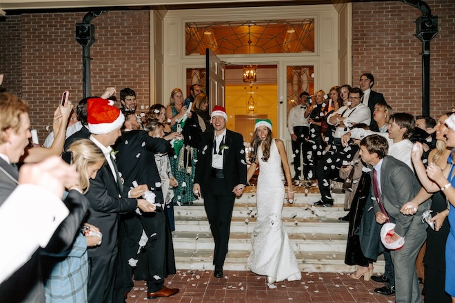 The bride and groom holding hands wearing Santa hats while guests throw confetti. 