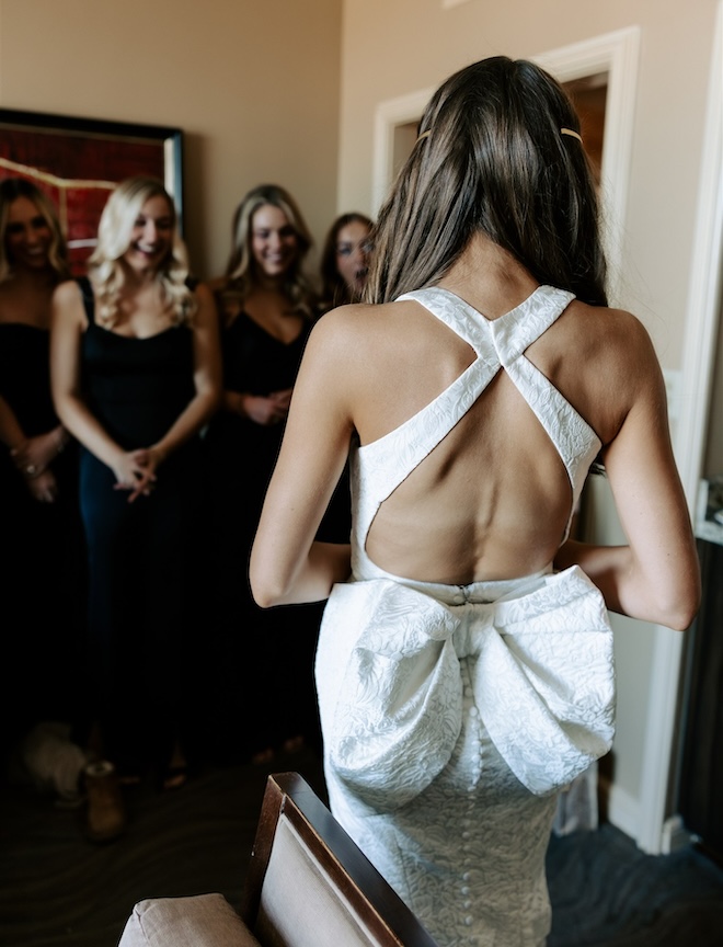 The back of the bride's wedding dress with a bow detail, with the bridesmaids smiling at the bride. 