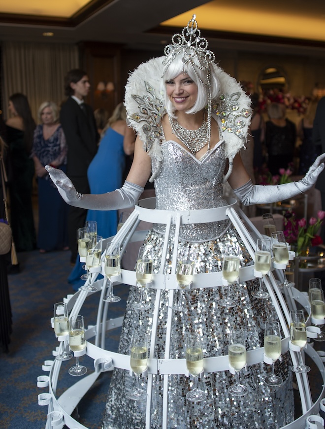 A woman wearing a silver sparkly dress that holds glasses of champagne. 