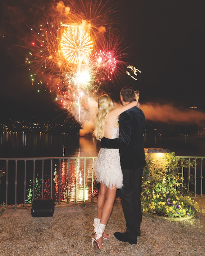 A bride and groom hugging while looking at a fireworks show over the water. 