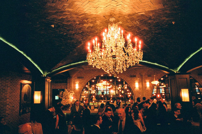 Low lighting chandelier in reception venue with brick ceilings and walls with guests around, setting the mood for their elegant autumn wedding.