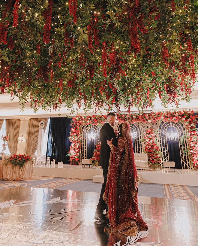 The bride and groom dancing under a canopy of greenery and red florals admiring their Beauty and the Beast-inspired decor. 