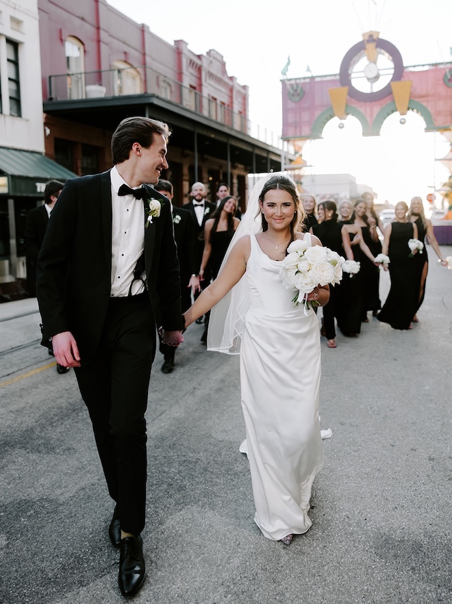 The bride and groom hold hands as they walk down the Galveston Strand as their wedding party follows. 