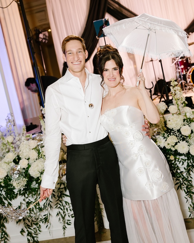 The bride and groom smiling while holding a white umbrella and streamers. 