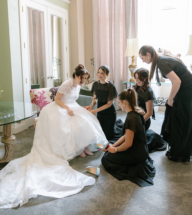 The bride laughing with her bridesmaids as they help put on her blue heels. 