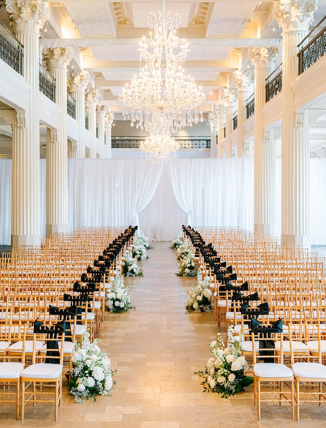 The aisle lined with white florals and chairs with black bows. 