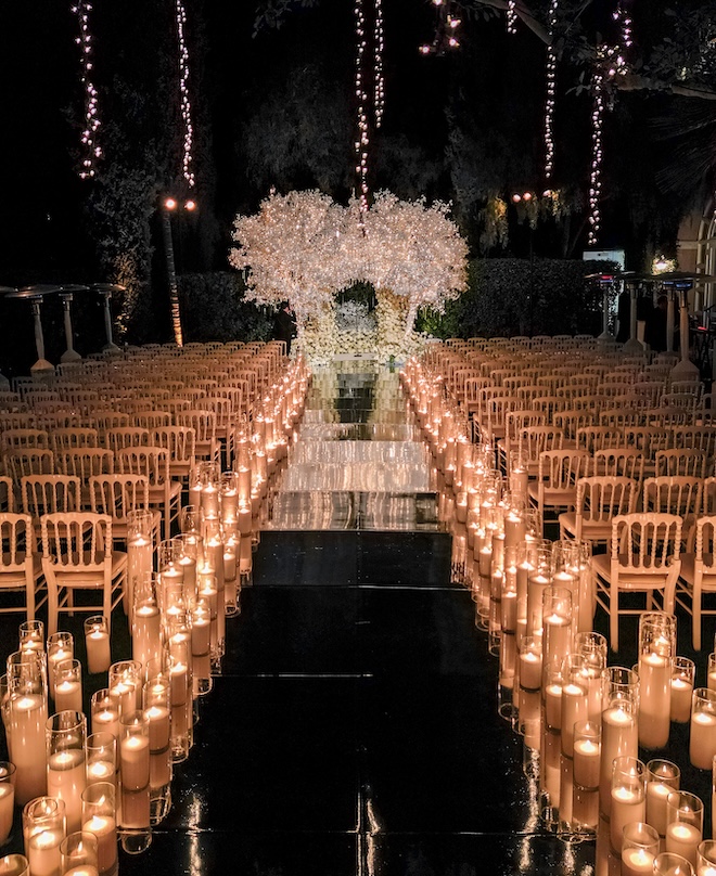 A ceremony decorated with candles down the aisle and a large floral installation at the altar with lights. 