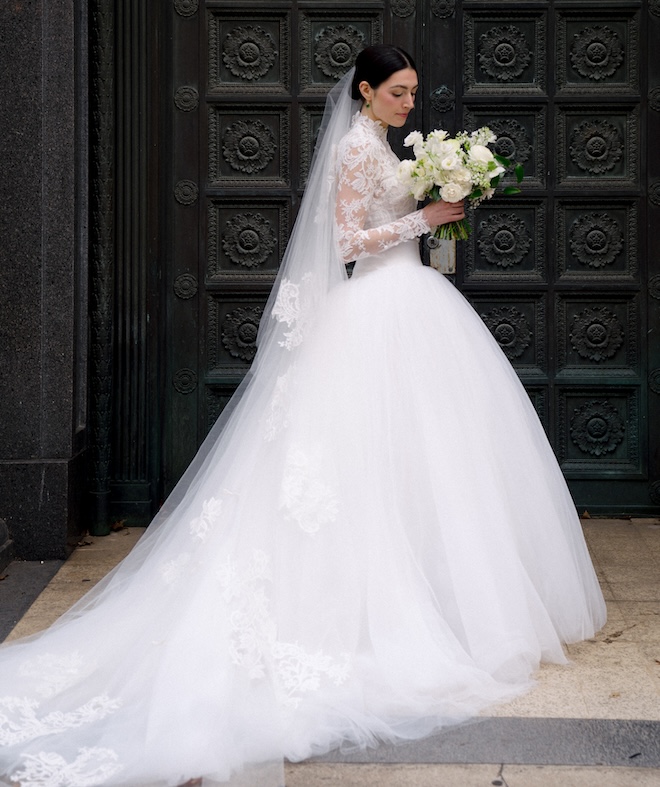 A bride wearing a lace-sleeved wedding gown with a full skirt and cathedral-length veil. 