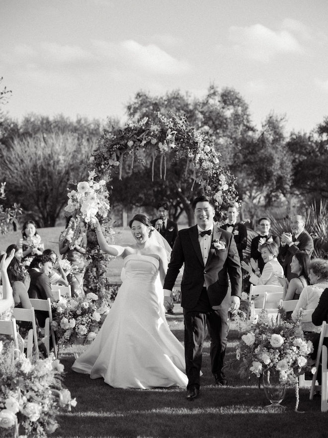 Black and white photo of the newlyweds walking down the aisle smiling and bride cheering with bouquet in the air.