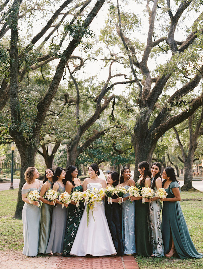Bride and bridesmaids outside by trees holding their yellow and white bouquets.