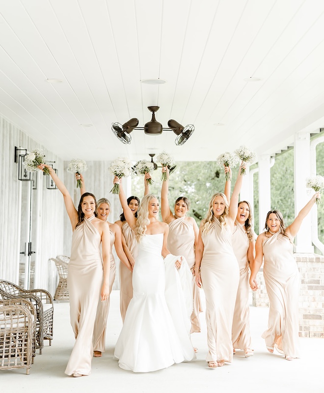 The bride and her bridesmaids walk on the porch of their wedding venue holding up their bouquets in celebration.