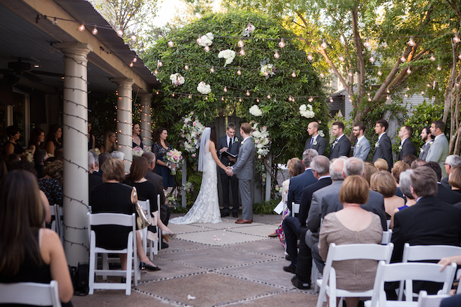 Bride and groom at outdoor altar with string lights and lush greenery.