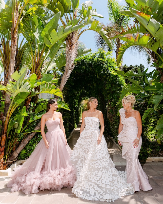 The bride stands next to her mother and sister in front of a canopy of greenery. 
