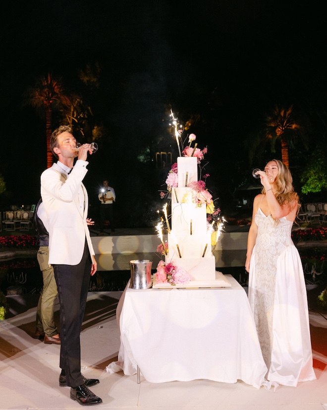 The bride and groom sip Champagne next to their five-tier white wedding cake. 