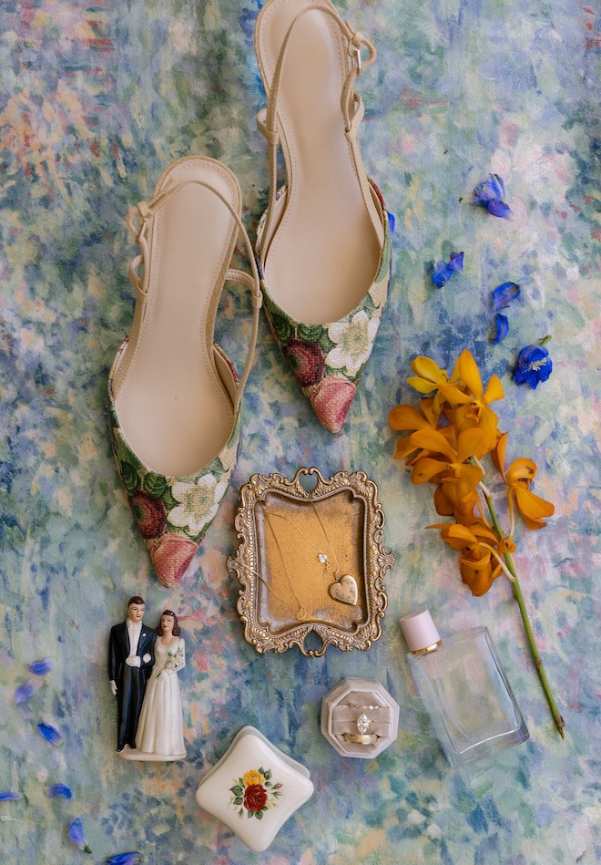 A flatlay on a watercolor background with floral shoes, bride and groom figurine, jewelry and flowers. 