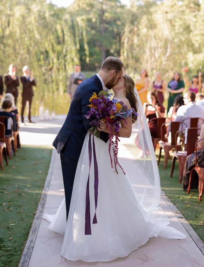 A bride and groom kissing down the aisle with the bride holding a jewel-toned bouquet. 