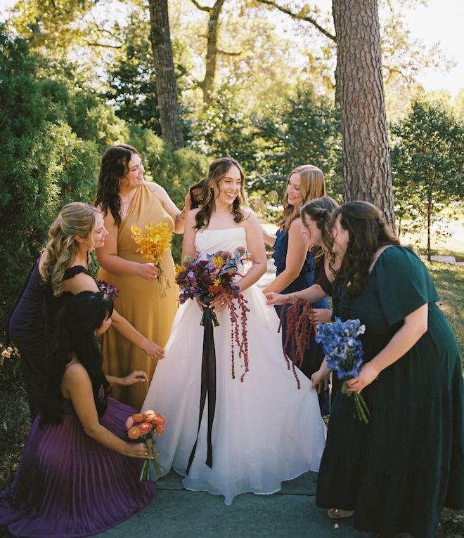 The bridesmaids helping the bride with her dress while all wearing mismatched dresses. 