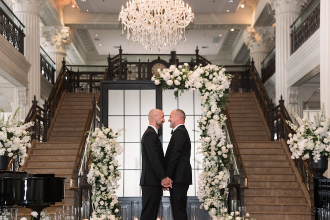The two grooms hold hands at the alter at their sophisticated black tie wedding.