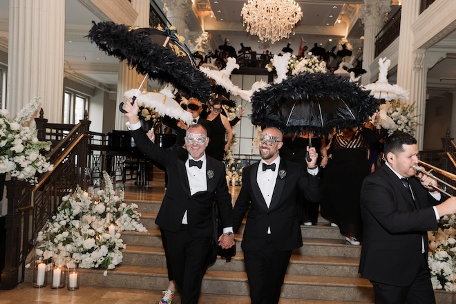 The two groom's hold hands as they walk down the grand stair case of the Corinthian Houston during their second-line parade.