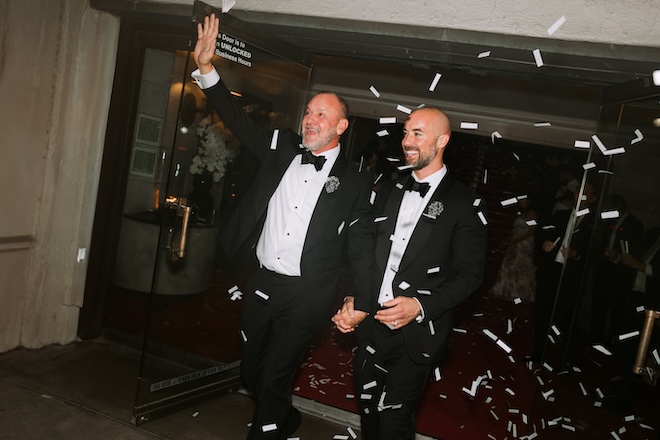 The couple walks out of their reception holding hands as streamers float in the air.