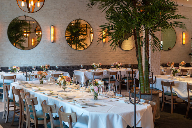 Reception tables with white linen and colorful small bouquets with a palm tree in between the tables, along with circular mirrors and dim lighting on the walls.