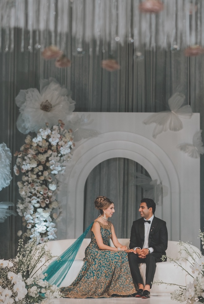 The couple sit on a white couch on a stage at their wedding reception. 