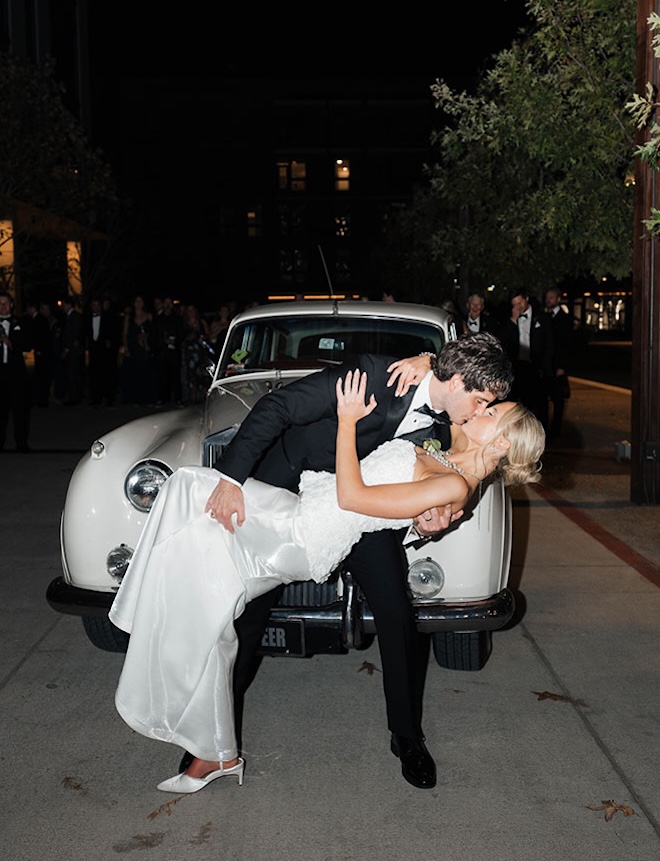 The couple share a kiss in front of a vintage white car. 