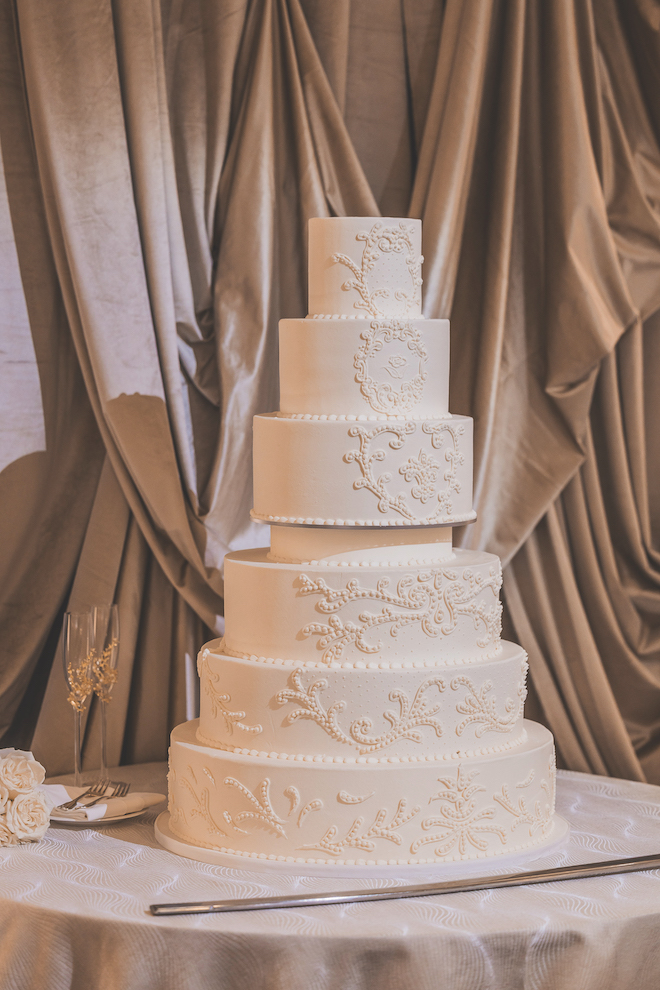 Close-up of six-tier wedding cake with textured embroidery in front of gold hued drapes.