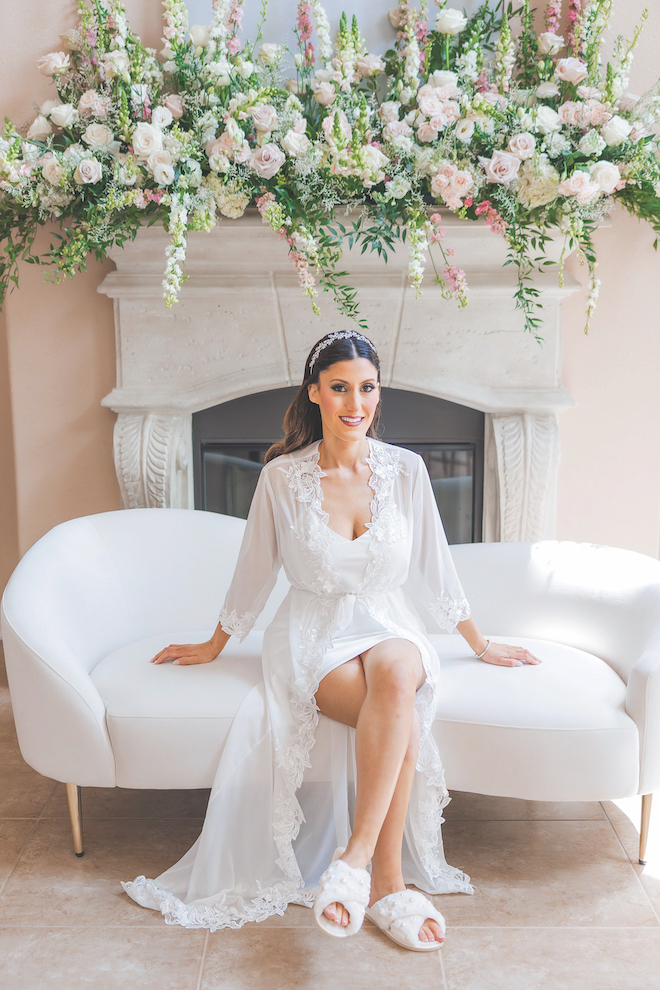 Bride in get ready outfit sitting on white couch in front of florals getting ready for her glamorous gold wedding at Houstonian.