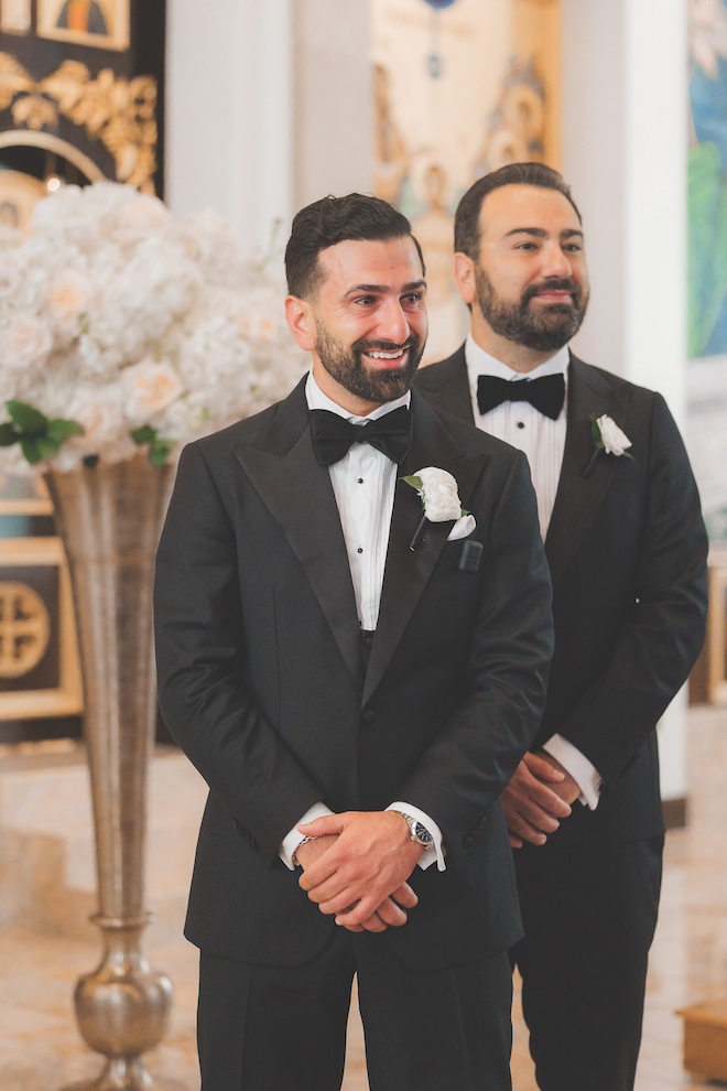 Groom standing at the aisle watching the bride walk down with tears in his eyes in front of elegant bouquet.