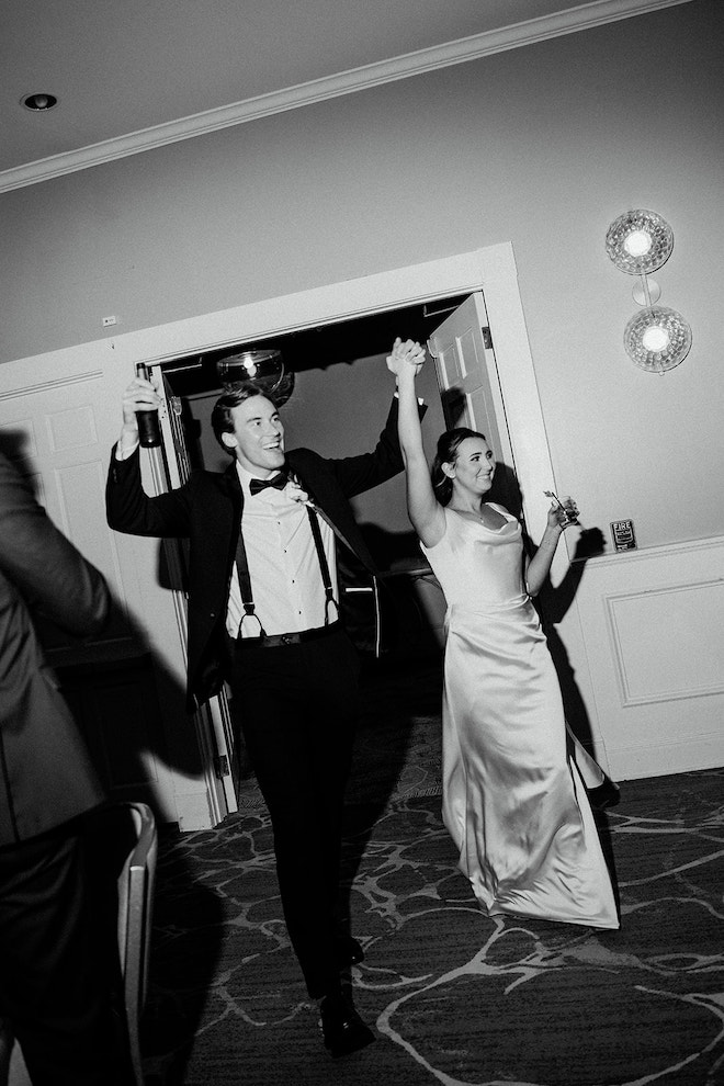 Black and white photo of bride and groom holding hands in the air entering the reception.