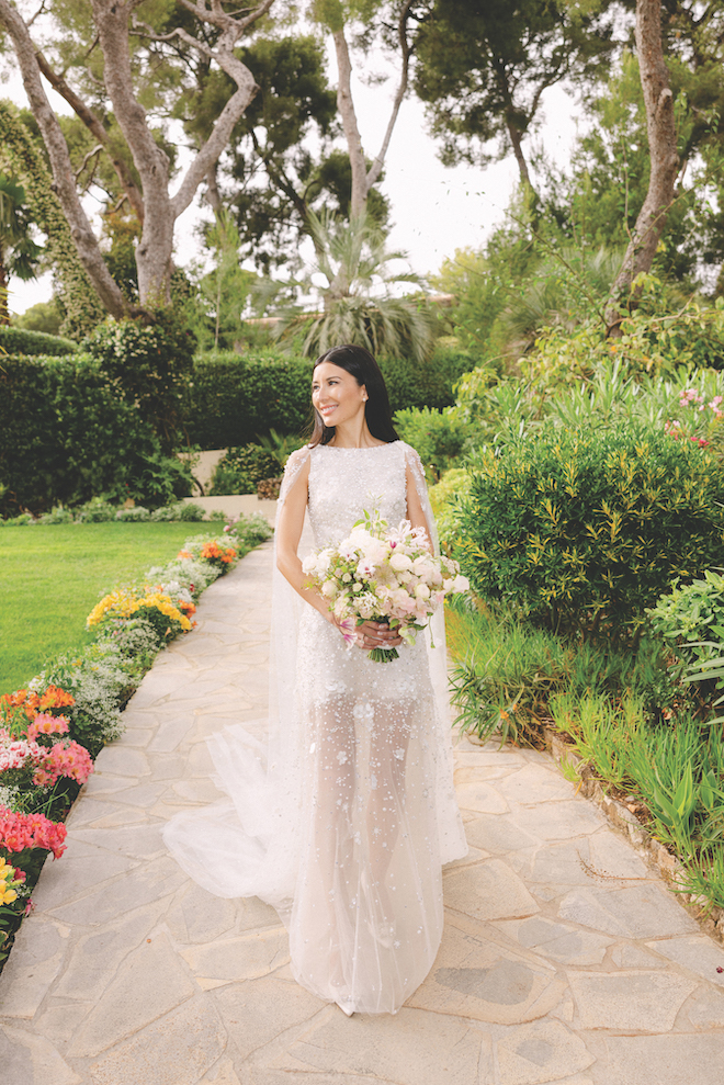 Bride standing on walkway lined with colorful flowers surrounded by greenery and holding white bouquet before walking down the aisle.