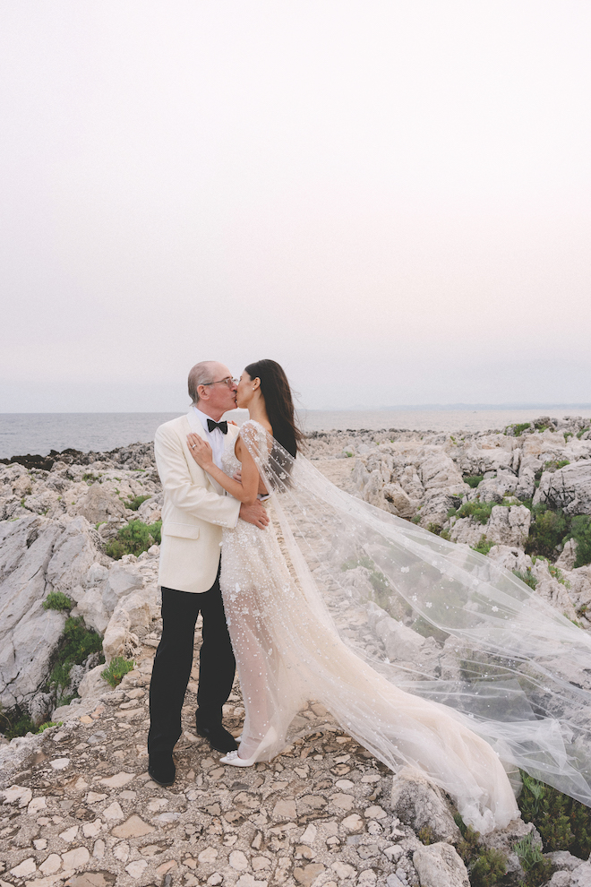 Newlyweds kissing on open rock with an ocean view as the bride's sheer dress and cape flowing in the wind.