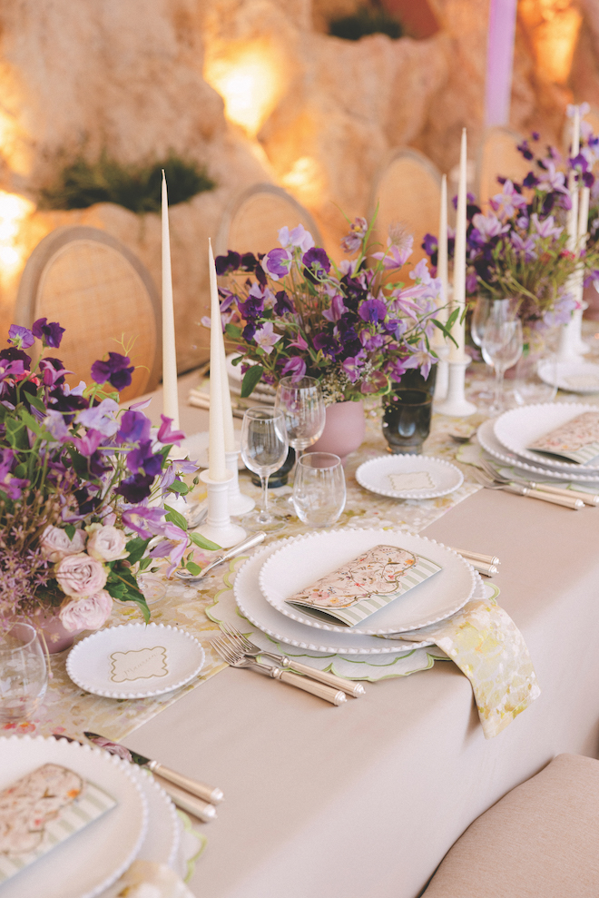 Purple flowers lining down the long reception table with white candles and floral detailed placemats and white table settings.