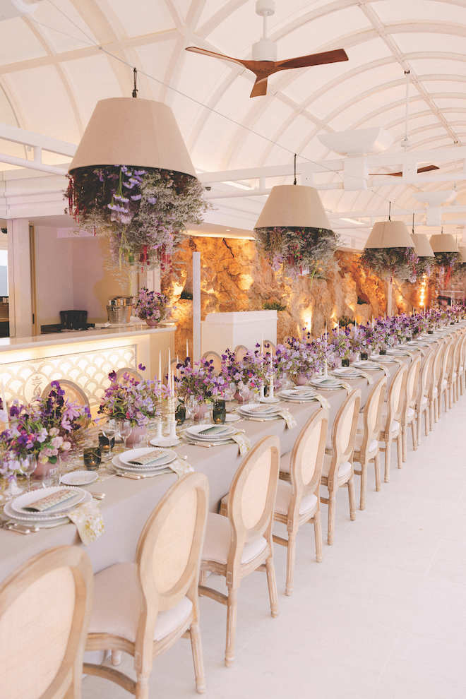 Long reception table with table setting details and purple flowers lining down the table with florals hanging out of chandliers.