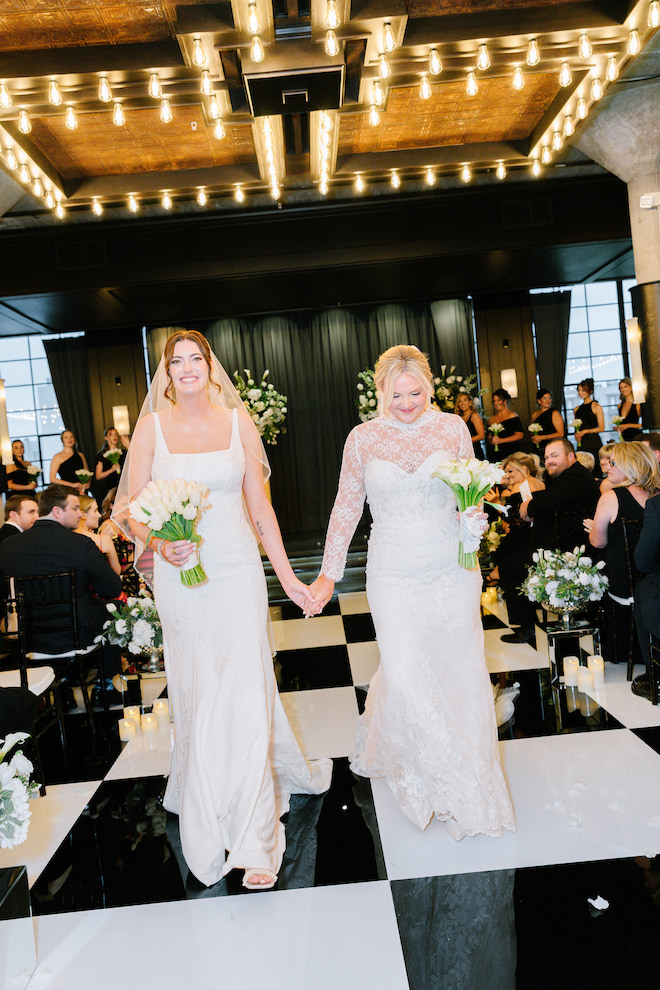 Newlyweds walking down the black and white checkered aisle holding hands and white bouquets for stunning spring wedding with white blooms.