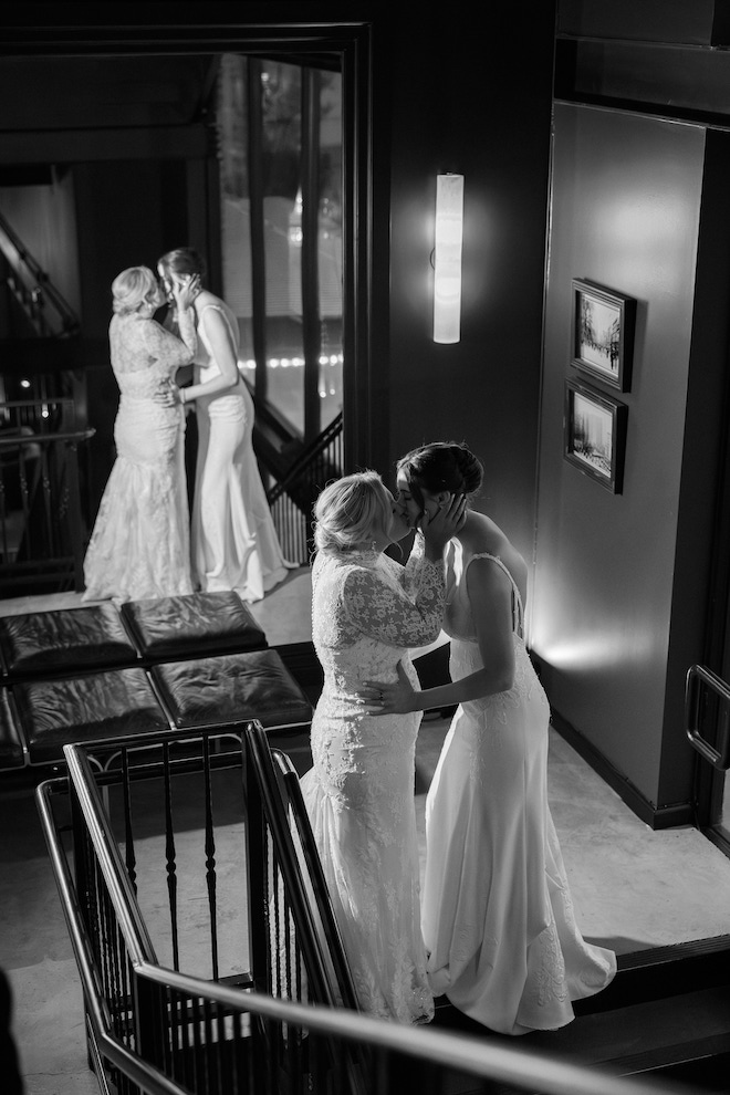 Black and white photo of the two brides kissing in stairwell in front of mirror.
