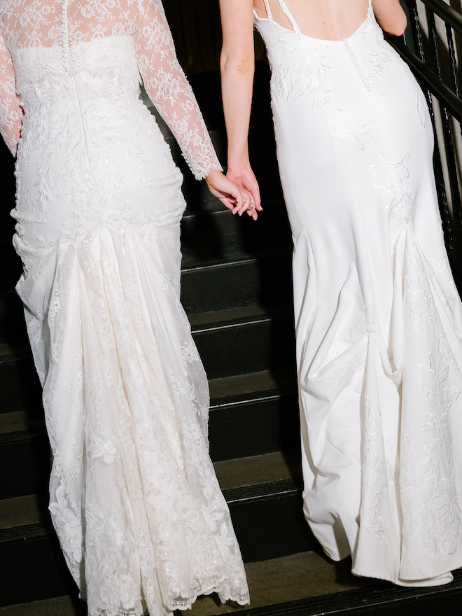 Behind photo of two brides holding hands going up the stairs, showing the backs of their dresses.