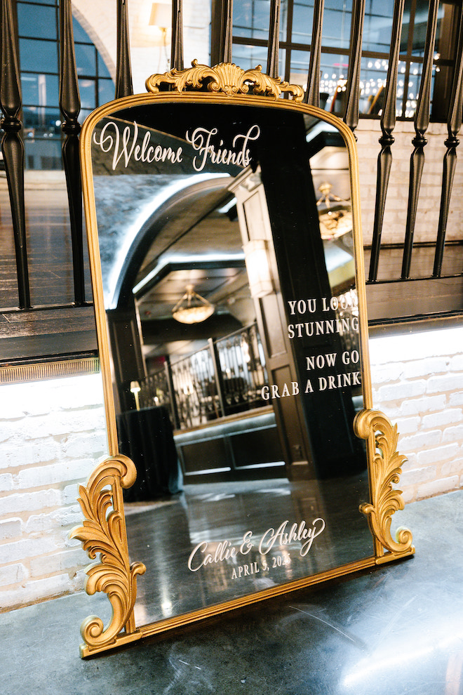 Gold trimmed mirror that reads, "Welcome Friends, You Look Stunning, Now Go Grab A Drink" for guests at cocktail hour.