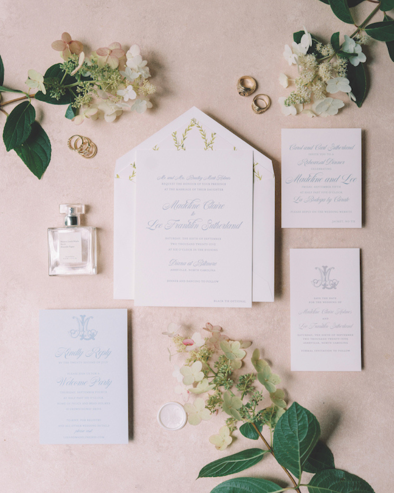 Stationery with white and blue details surrounded by greenery and perfume for unique wedding invitation ideas.