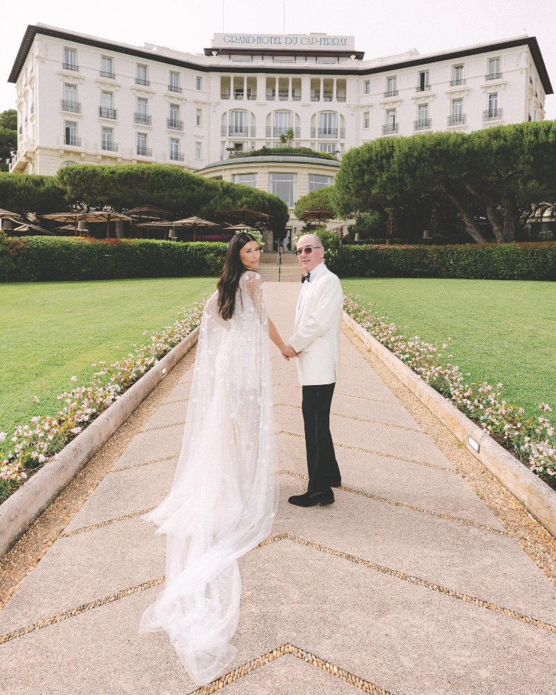 Bride and groom walking towards hotel venue with spring florals lined down the walkway while they look back at the camera and hold hands.