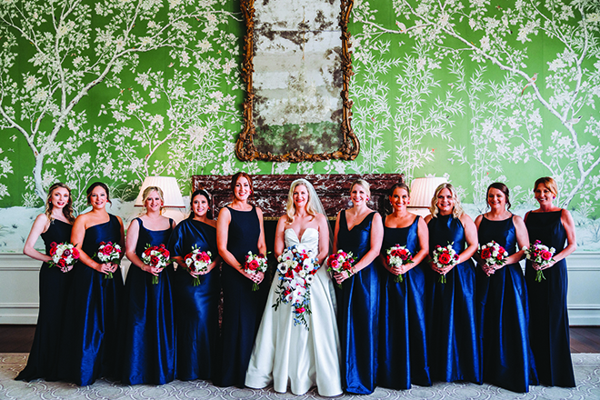 Bride standing with bridesmaids in dark blue dresses holding multi-colored bouquets in front of green wall with white details.