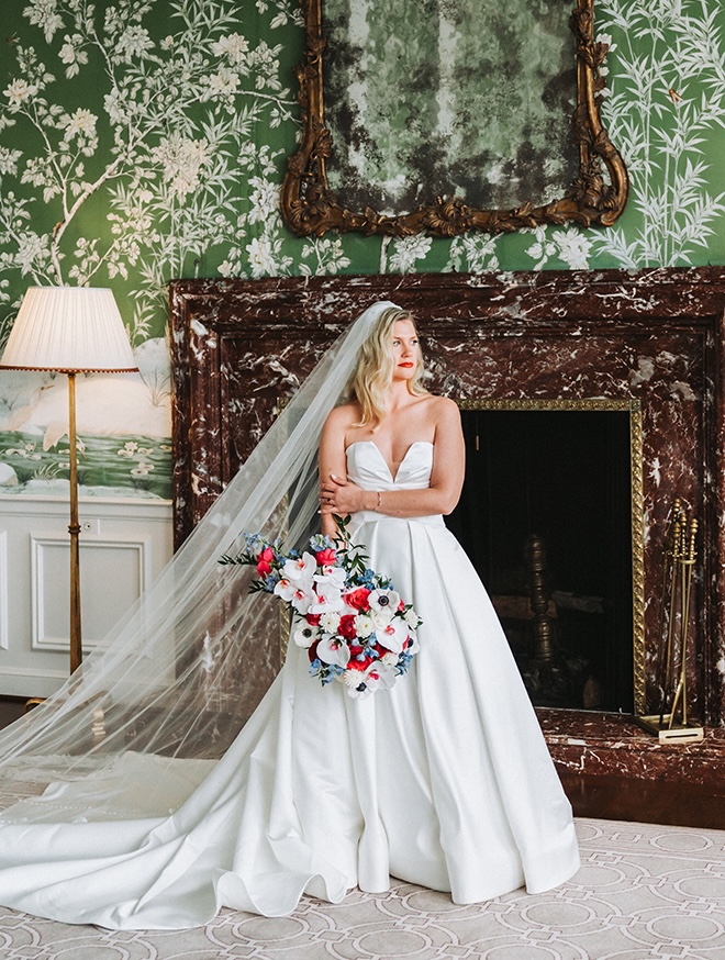 Bride in stunning gown holding multi-colored bouquet with long veil in front of vintage fireplace.