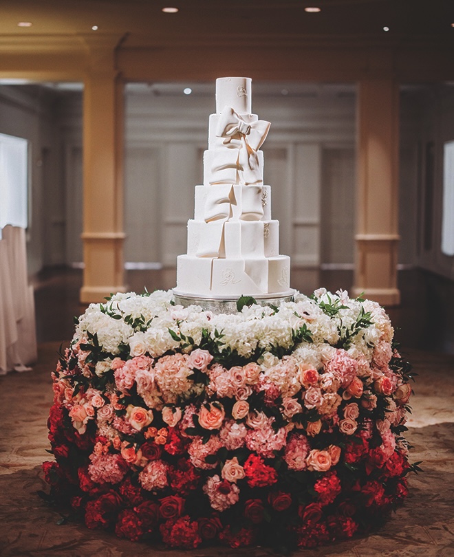 White six-tier wedding cake with edible bow on top of a bouquet table filled with red, pink, and white roses.