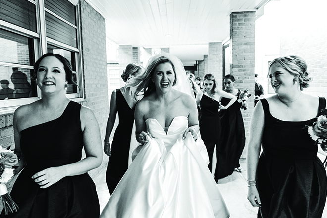 Black and white photo of bride walking in between bridesmaids smiling getting ready to walk down the aisle.